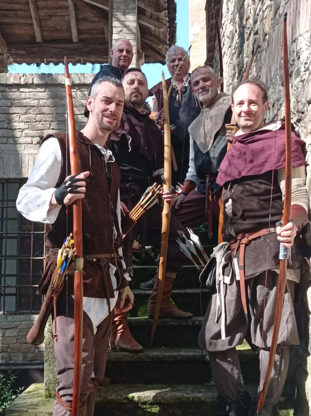 Persone in costume medievale con archi e frecce su una scala in pietra, davanti a edifici storici. I volti sono oscurati.