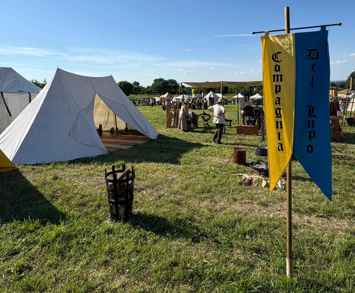 Campo di rievocazione medievale con tende bianche, persone in costume d'epoca e uno striscione blu e giallo con la scritta “Compagnia del Lupo” su un prato.