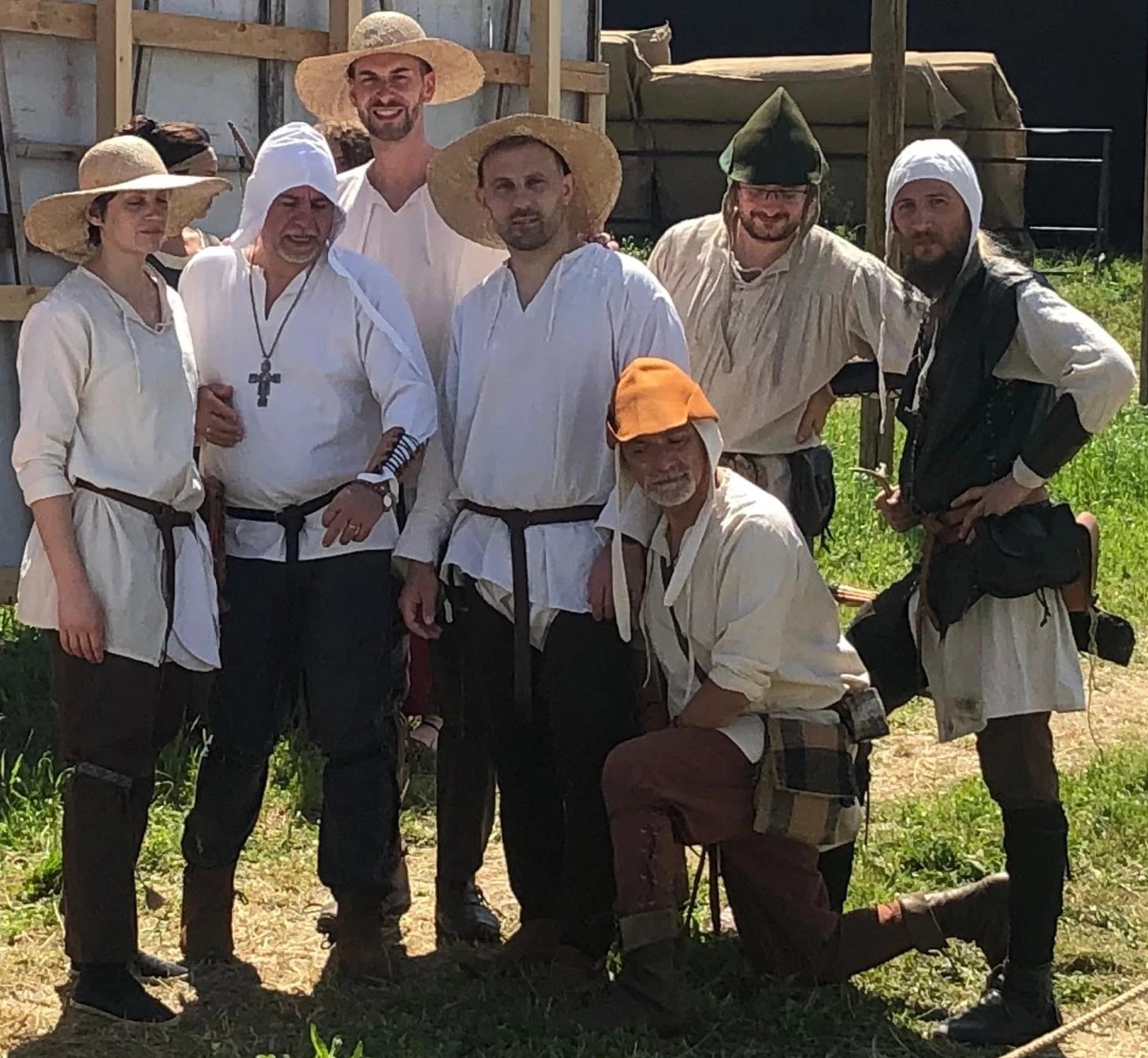 Un gruppo di sei persone vestite con abiti storici e cappelli di paglia posano insieme in un ambiente erboso all'aperto, sorridendo allegramente.