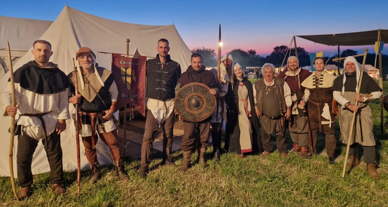 Un gruppo di sette individui in abiti medievali, in piedi davanti a una grande tenda bianca al tramonto, con in mano vari oggetti di scena e armi.