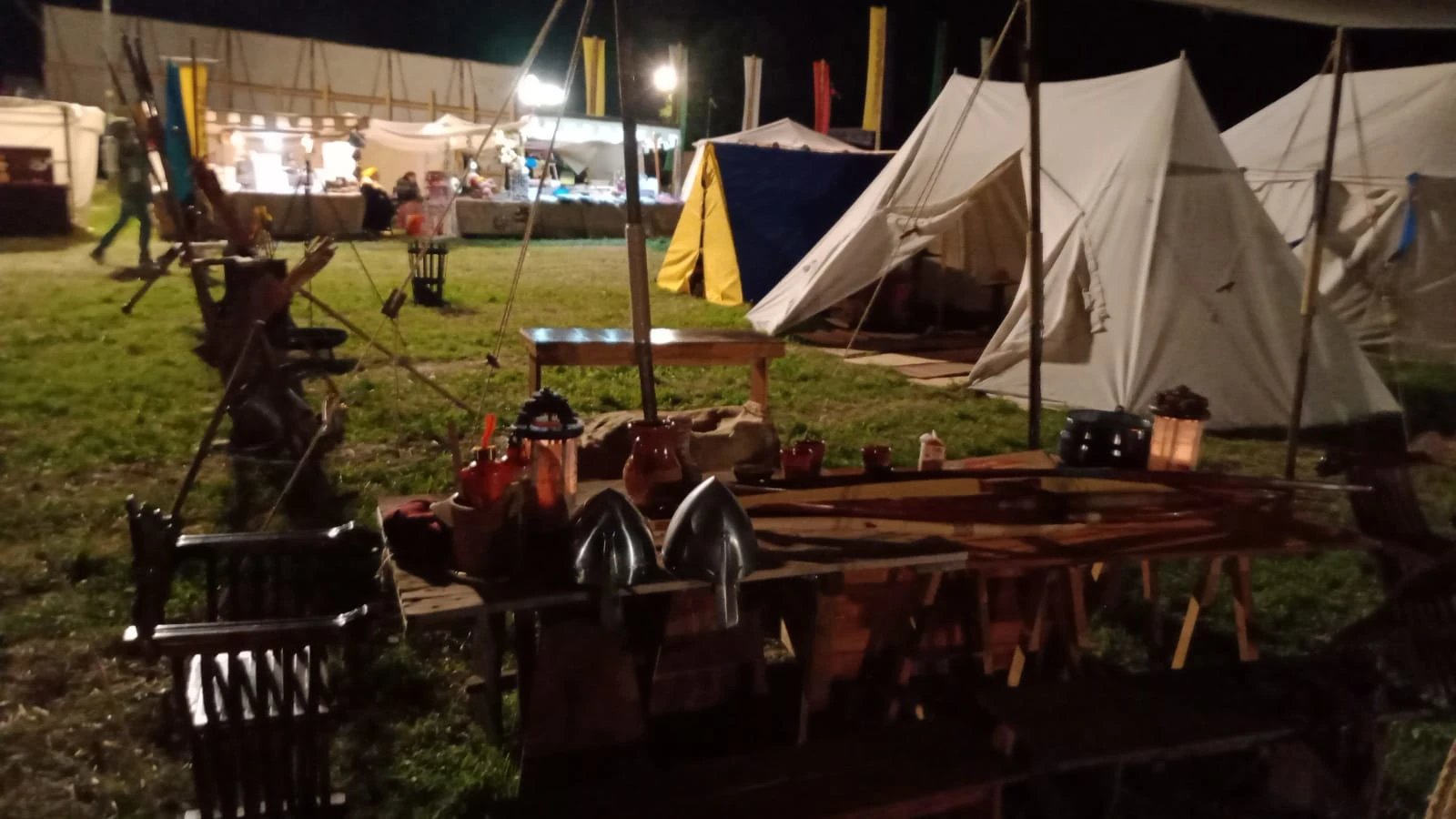Accampamento notturno all'aperto a tema medievale con tende, tavoli di legno, elmi e vari oggetti di scena; sullo sfondo si vedono delle persone vicino a una tenda illuminata.