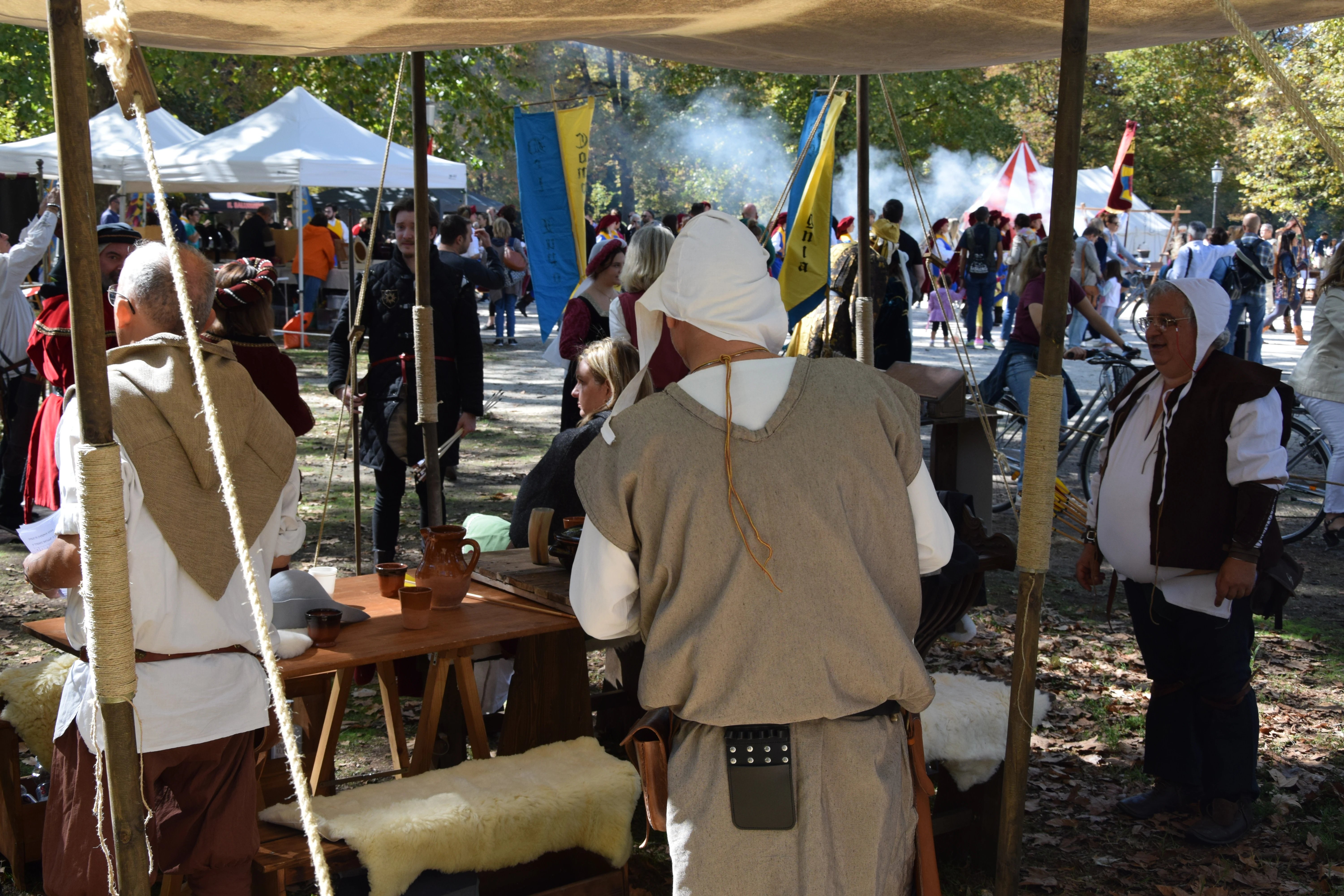 Scena di festa medievale con persone in costumi d'epoca, tende, stendardi e fumo in un parco. Sono visibili tavoli di legno e vasellame.